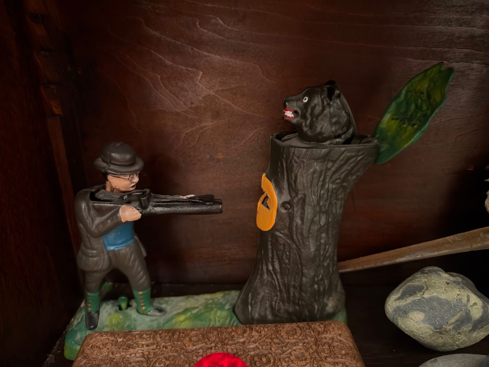 A painted mechanical bank showing a hunter in a bowler hat aiming at a black bear popping out of a tree stump