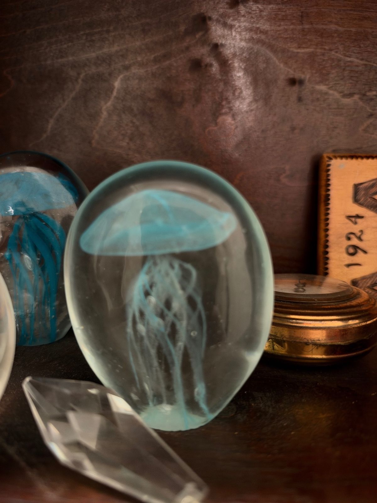 Two glass jellyfish paperweights, one pale blue and one deep turquoise, with a faceted crystal point in front and a round brass compass behind